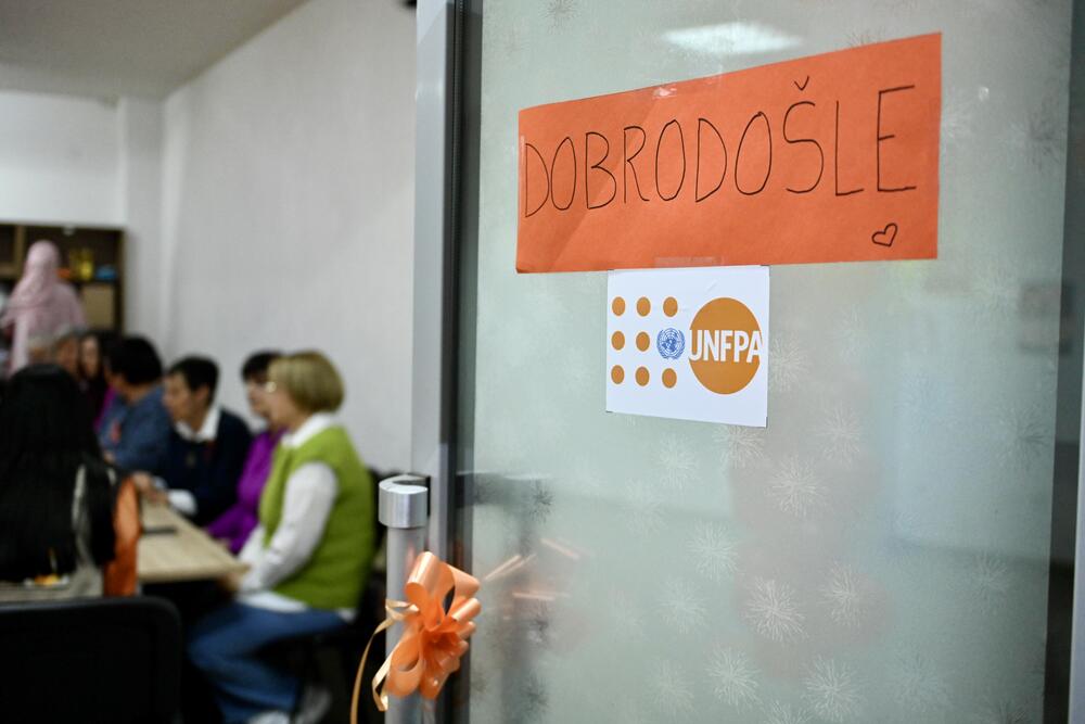 Vrata ukrašena narandžastom mašnom i natpisom "DOBRODOŠLE" te logotipom UNFPA. U pozadini grupa ljudi sjedi za stolom i učestvuje u diskusiji.