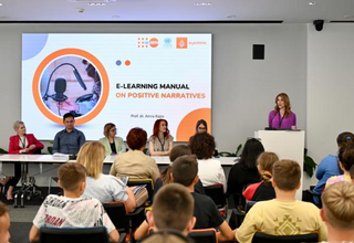 The image depicts a professional seminar with an audience of adults and children listening to a speaker at a podium, presenting 