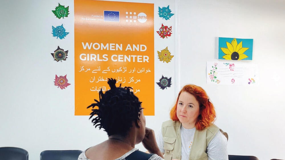 In the image, a dark-skinned woman, seated with her back to the camera, is talking to a white woman with red hair who is wearing a vest with the UNFPA symbol. Behind them is a large orange banner that reads "WOMEN AND GIRLS CENTER" and features the logos of the European Union and UNFPA. The wall is decorated with colorful paper leaves and drawings. The room appears to be a safe and supportive environment, likely within a temporary reception center.