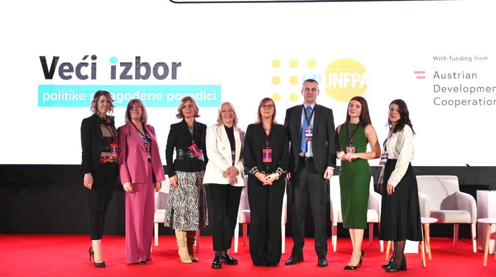 A group of eight people stands on a stage with a red carpet, posing for a photo in front of a large screen displaying the UNFPA logo and text that reads "Veći izbor" and "politike prilagođene porodici." The event is funded by the Austrian Development Cooperation. The group includes a mix of men and women, all dressed in professional attire, with some wearing badges around their necks. The atmosphere appears formal, with a focus on family-friendly policies, as indicated by the background text.