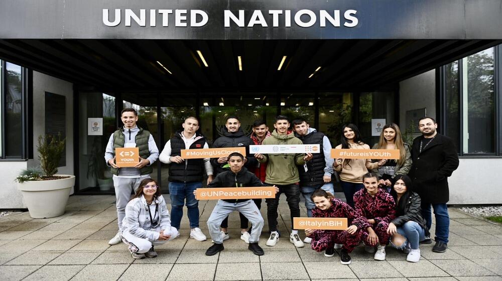 A group of young men and women pose outside the United Nations building in Bosnia and Herzegovina, holding signs with hashtags like "#MovingUsCloser," "#futureisYOUth," "#UNPeacebuilding," and "#ItalyinBiH," as part of the Y-Peer network's youth peacebuilding initiative.