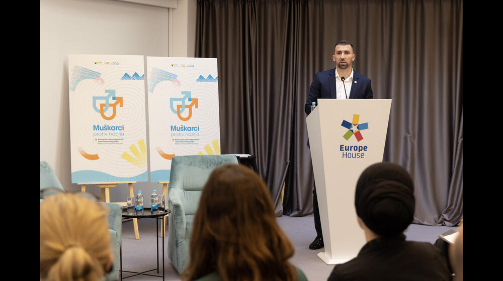 A speaker at the "Men Against Violence" event delivers a speech from a podium at Europe House, while the audience listens. Event banners in the background display the logo and name, promoting male engagement in preventing gender-based violence.