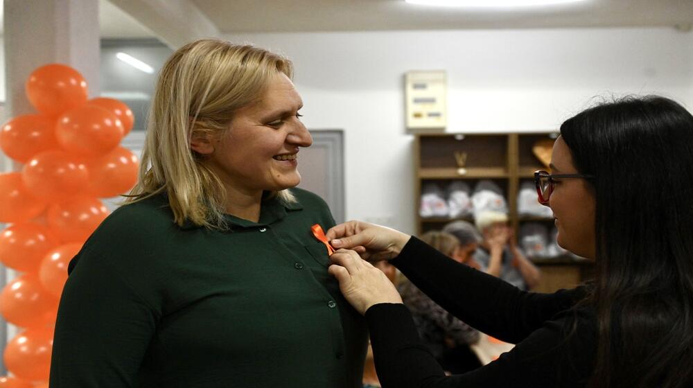 A woman with a smile receives an orange ribbon, which another woman pins to her shirt. In the background, balloons and other event participants are visible.