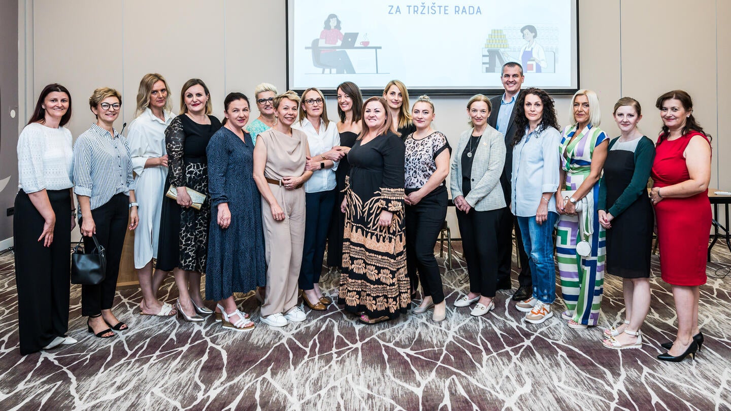 Grupa profesionalno obučenih žena i jedan muškarac stoje zajedno u konferencijskoj sali, pozirajući za grupnu fotografiju. Svi se smješkaju i gledaju u kameru. Pozadina prikazuje projektorski ekran sa naslovom 'OSPOSOBLJAVANJE DUGOTRAJNO NEZAPOSLENIH ŽENA ZA TRŽIŠTE RADA'.