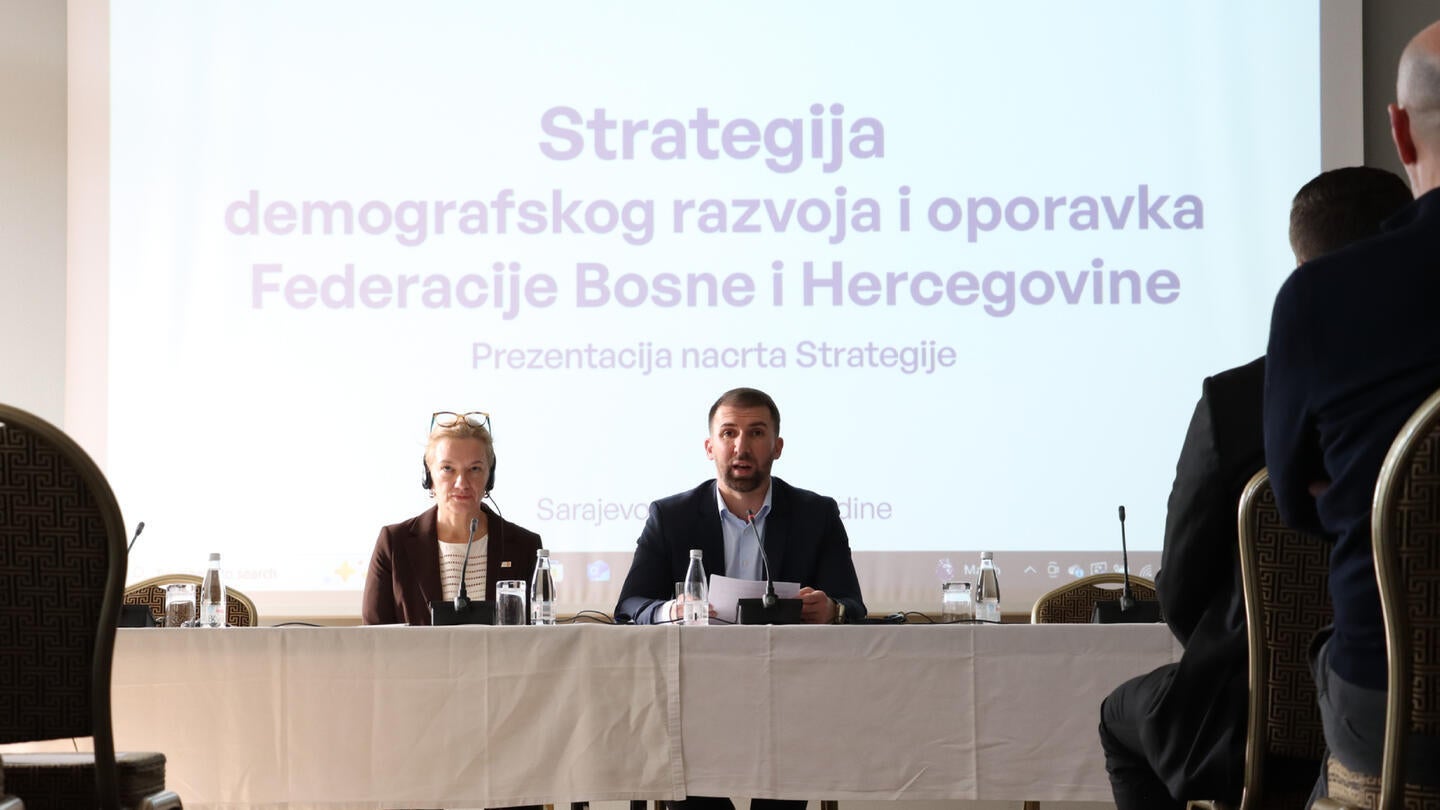 Prezentacija o "Strategiji demografskog razvoja i oporavka Federacije Bosne i Hercegovine". Muškarac u tamnom odijelu i žena u smeđem sakou sjede za stolom i govore u mikrofona. Iza njih na ekranu projektora prikazan je naziv strategije velikim ljubičastim slovima. Publika, obučena u formalnu odjeću, sjedi u redovima stolica i pažljivo sluša.