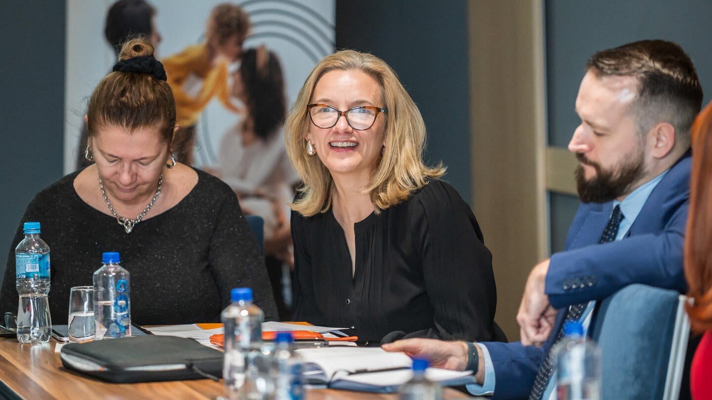 A woman with blonde hair, smiling, seated at a conference table during a panel discussion, accompanied by two other participants, a focused woman and a bearded man, in a modern meeting room with bottled water and notebooks on the table