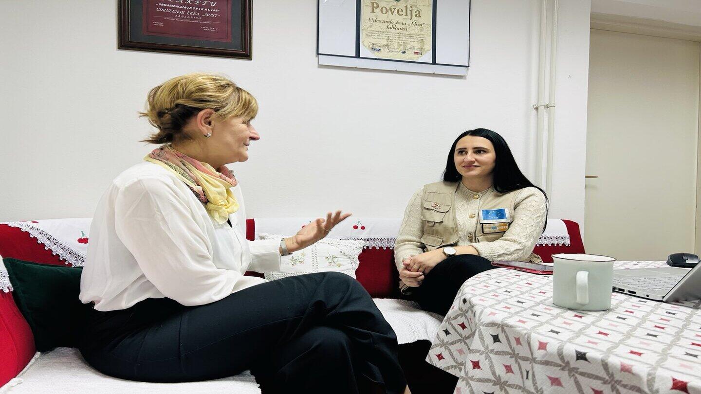 Two women sit on a couch in a warmly decorated room, engaged in conversation. The woman on the left, wearing a white blouse and a colorful scarf, gestures as she speaks, while the woman on the right, dressed in a beige vest with an EU-UNFPA badge, listens attentively. A table with a patterned tablecloth, a laptop, and a mug is in front of them. Behind them, framed certificates and embroidered cushions decorate the space.
