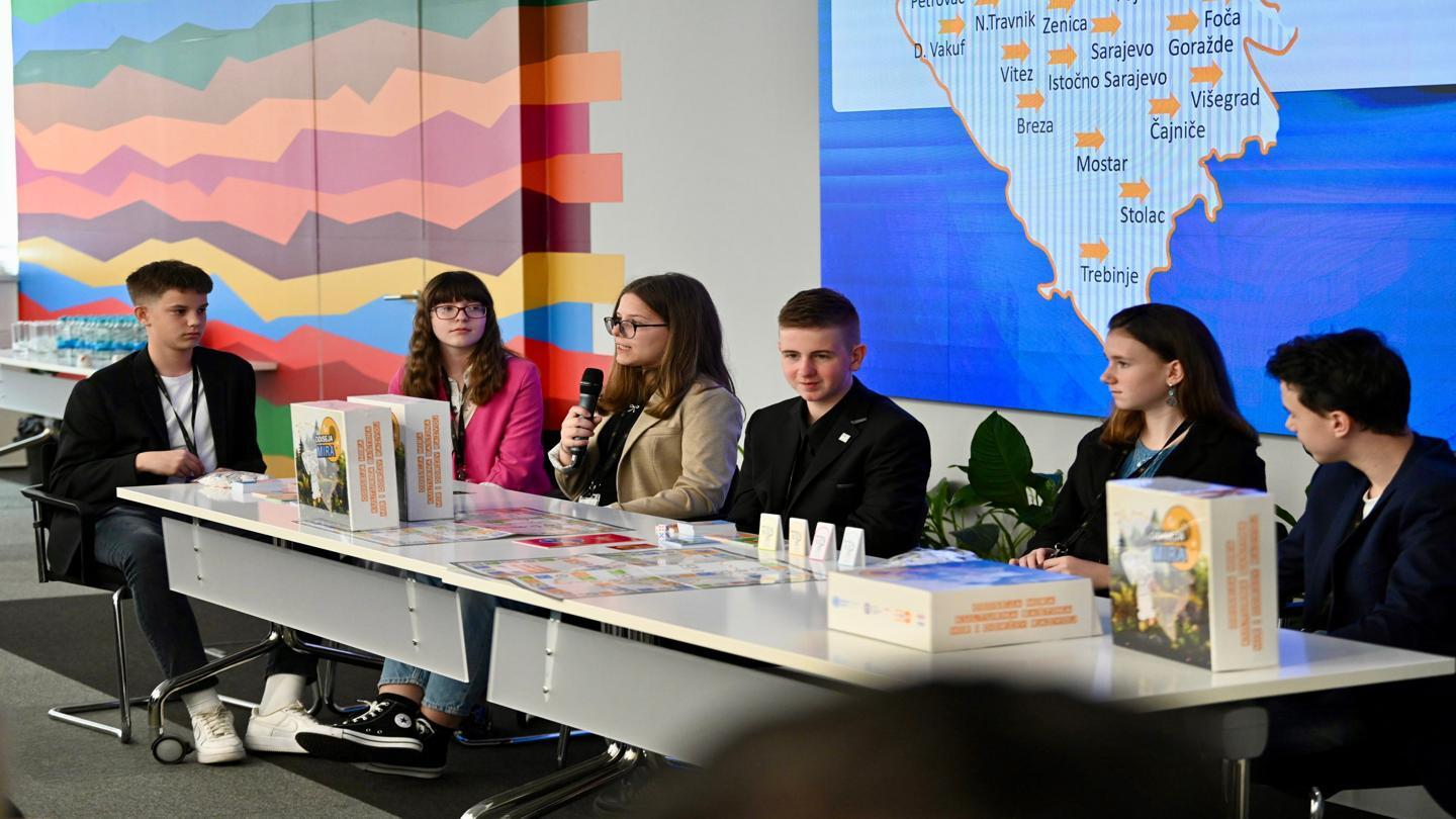 A group of high school students from across Bosnia and Herzegovina seated at a table presenting a board game at the UN House in Sarajevo. Behind them is a map showing participating cities, and the table displays game boxes and promotional materials.