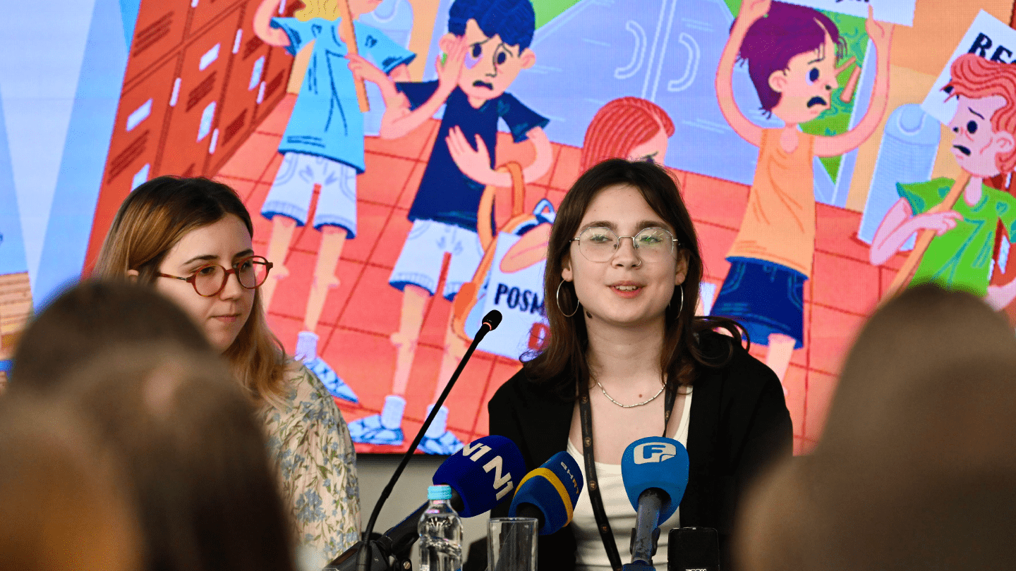 Two young women speak at a press event with illustrated anti-violence posters behind them.